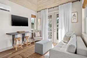 A modern living room with a sofa, ottoman, and bar stools. A flat-screen TV is mounted on the wall, and there is a window with curtains. - Charming Carriage House, Prime Location- Walk to Rosemary and 30A (Inlet Beach)