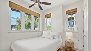 Una camera da letto con un letto, una ventola a soffitto, una finestra vista palme e un piccolo tavolo con una lampada.