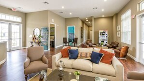 Amplia sala de estar con un sofá color beige, sillones y una mesa de centro. Varias puertas dan acceso a otras habitaciones, y se observa una máquina expendedora al fondo.