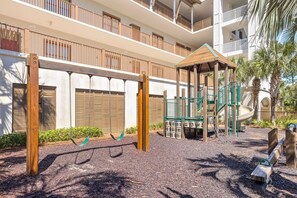 Un terrain de jeux avec des balançoires et un toboggan, entouré d’un bâtiment doté de balcons et d’un espace de jeux couvert.