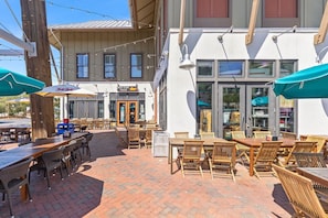 Un espace extérieur aménagé en terrasse avec des tables et des chaises en bois, un restaurant dont l’enseigne indique qu’il sert des tacos aux poissons, et un bâtiment doté de grandes fenêtres et d’une façade blanche.