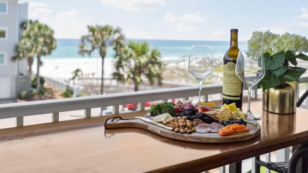 Ein Holztisch mit einer Wurstplatte, einer Caymus-Weinflasche und einem Weinglas auf einem Balkon mit Blick auf einen Strand.