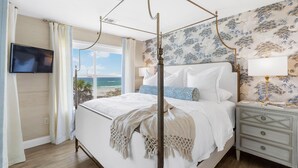 Ein Schlafzimmer mit Himmelbett, Wand-Fernseher, Nachttisch mit Lampe und Meerblick durch große Fenster.