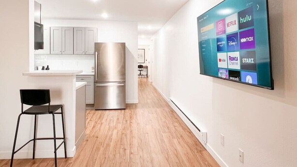 Una cocina moderna con armarios blancos, un refrigerador de acero inoxidable y una barra con una silla negra. Un televisor de pantalla plana montado en la pared muestra diferentes servicios de transmisión.
