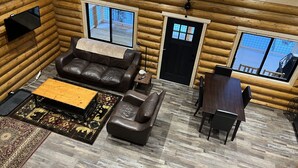 Ein Interieur einer Blockhütte mit einem Ledersofa, einem Holztisch, einem Esstisch mit Stühlen und großen Fenstern mit Blick auf eine verschneite Landschaft.