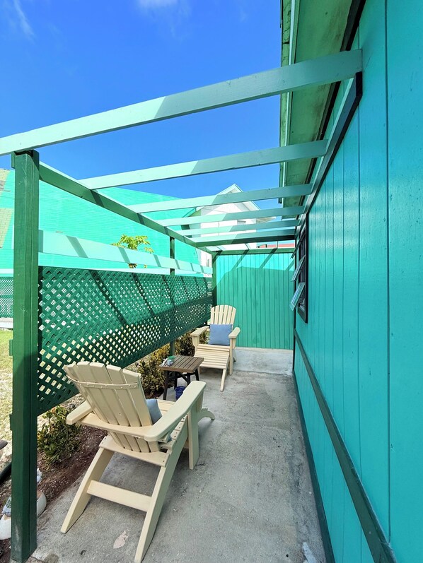 Een overdekt terras met twee stoelen, een tafel, een groen hek en een gebouw met blauwgroene bekleding.