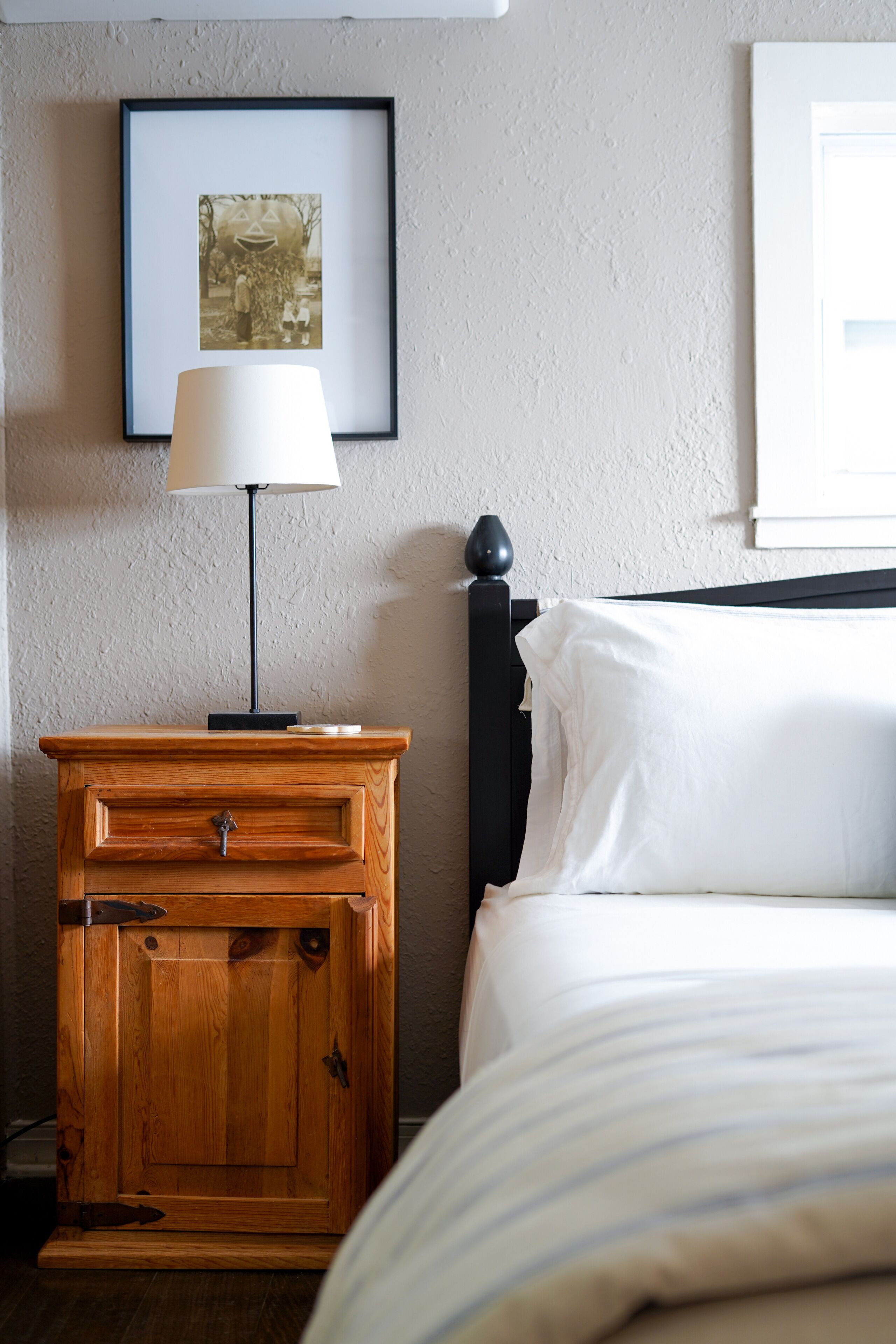 Een net opgemaakt bed met witte lakens, een houten nachtkastje met een lamp, en een ingelijst beeld aan de muur.