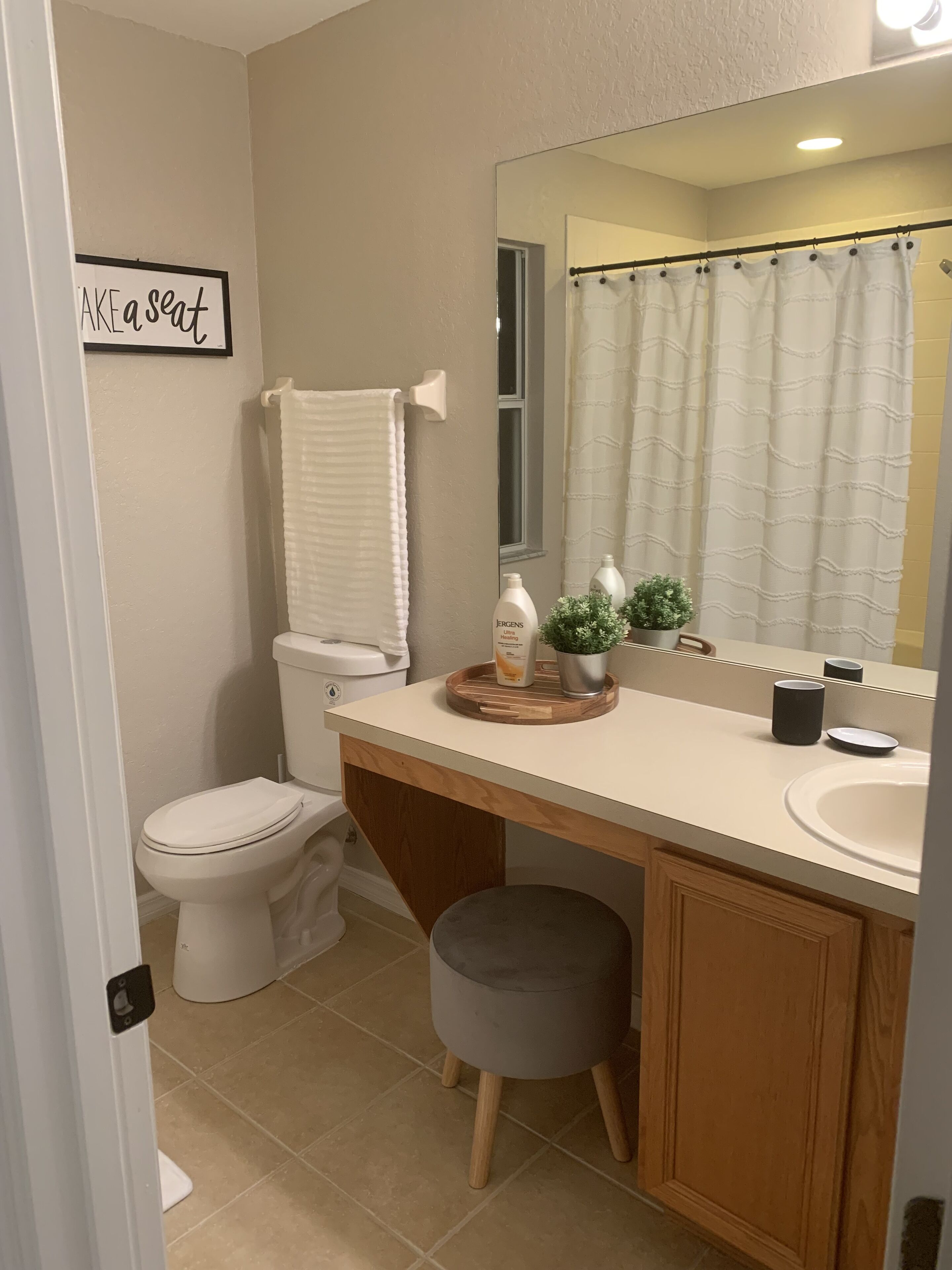 A bathroom with a toilet, sink, and mirror. There is a sign on the wall that says "Take a seat."