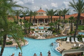 Un resort con una amplia piscina central, palmeras y un edificio de techo con tejas rojas.