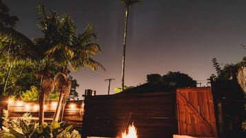 A nighttime outdoor setting with a fire pit, palm trees, and a wooden fence.