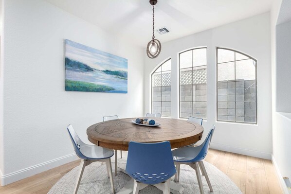 A dining area with a round wooden table, blue chairs, a painting on the wall, and large windows with a view.