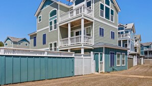A multi-story residential building with a balcony on the second floor, a fenced yard, and a clear blue sky. - Coastal Luxury | Hot Tub • Game Room • Pool (Corolla)