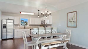 A modern kitchen with white cabinetry, a stainless steel refrigerator, a built-in microwave, and a dining area with a wooden table and white chairs. - Coastal Luxury | Hot Tub • Game Room • Pool (Corolla)
