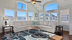 A spacious living room with a white sectional sofa, a coffee table, and a patterned rug. Large windows offer views of the neighborhood and a church steeple. - Coastal Luxury | Hot Tub • Game Room • Pool (Corolla)