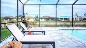 A poolside area with lounge chairs, a table with fruit, and a view of houses across a body of water. - Paradiso - Modern Style Vacation Villa (Cape Coral)