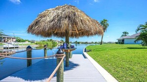 A thatched-roof structure on a wooden dock with a clear view of a water body and a grassy area.