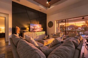 A modern living room with a large sofa, a flat-screen TV, and a view of the sunset through large windows.
