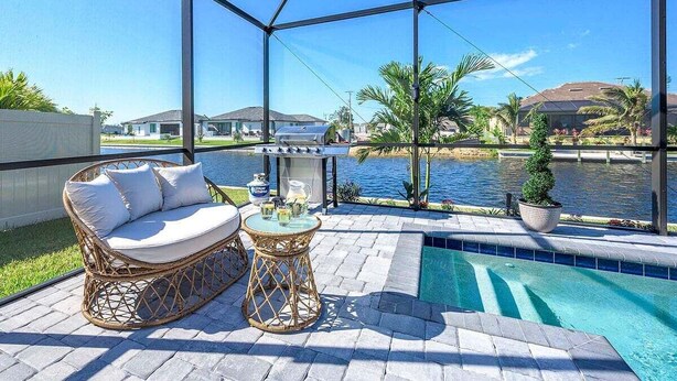 A covered outdoor area with a pool, wicker furniture, and a view of a body of water and houses.