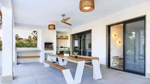 A modern outdoor patio with a wooden table and benches, a ceiling fan, and a glass door leading to a balcony. - 5 Star Villa Magnolia – Luxury Lakefront Retreat (Algarve)