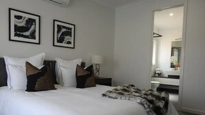 A modern hotel room with a large bed, white linens, and decorative pillows. There are two framed abstract paintings on the wall, a bedside lamp, and a view into the bathroom through an open door.