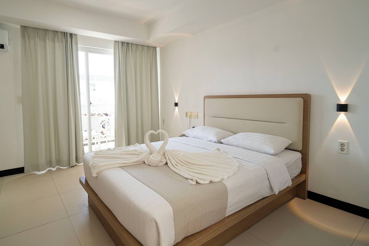 Une chambre d’hôtel avec un lit, des rideaux, une lampe fixée au mur et une vue par la fenêtre.