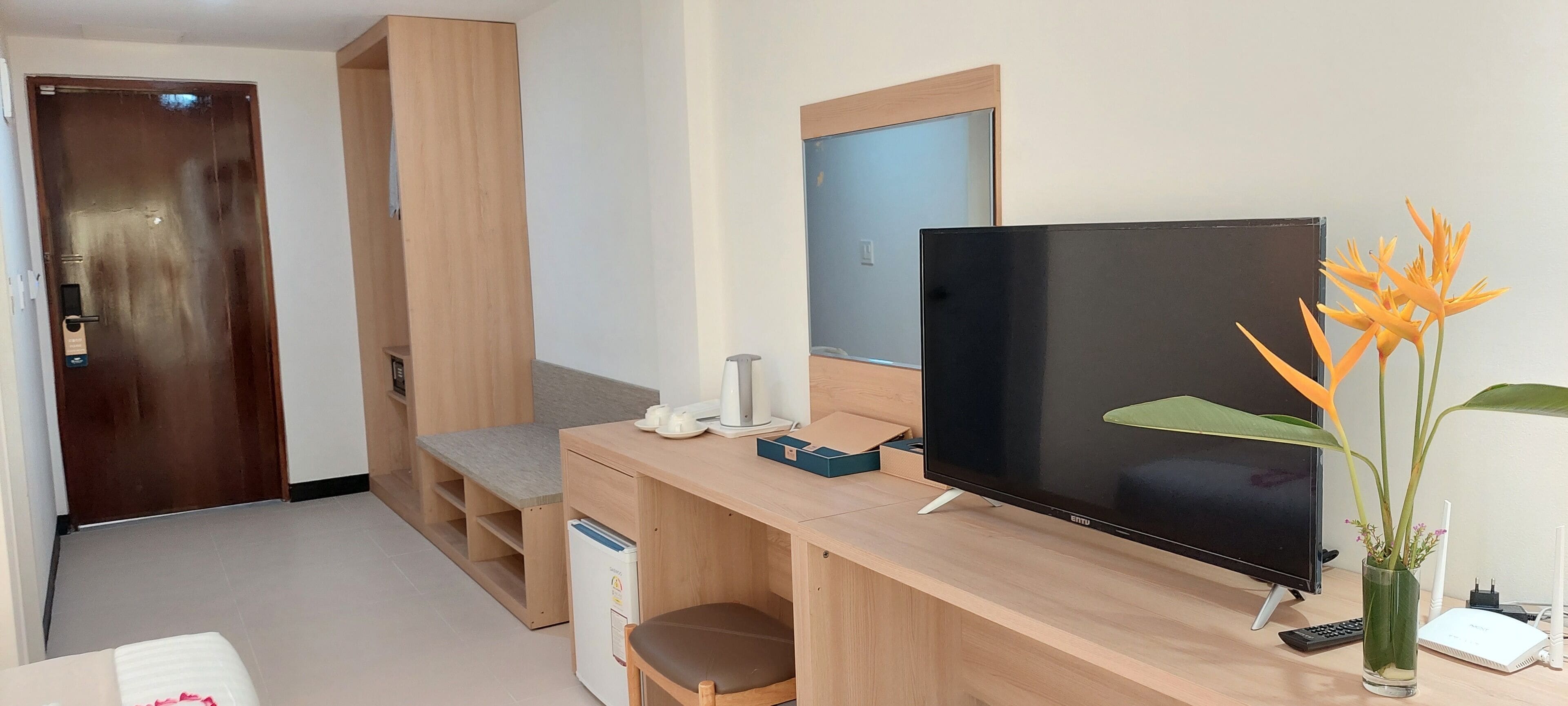 Habitación de hotel compacta con escritorio de madera, televisor de pantalla plana, una pequeña zona de estar y un jarrón con una sola flor.