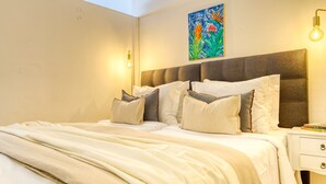 Un letto rifatto con precisione, con una testiera grigia, cuscini bianchi e grigi e una coperta beige. È presente un comodino con una lampada e un libro.