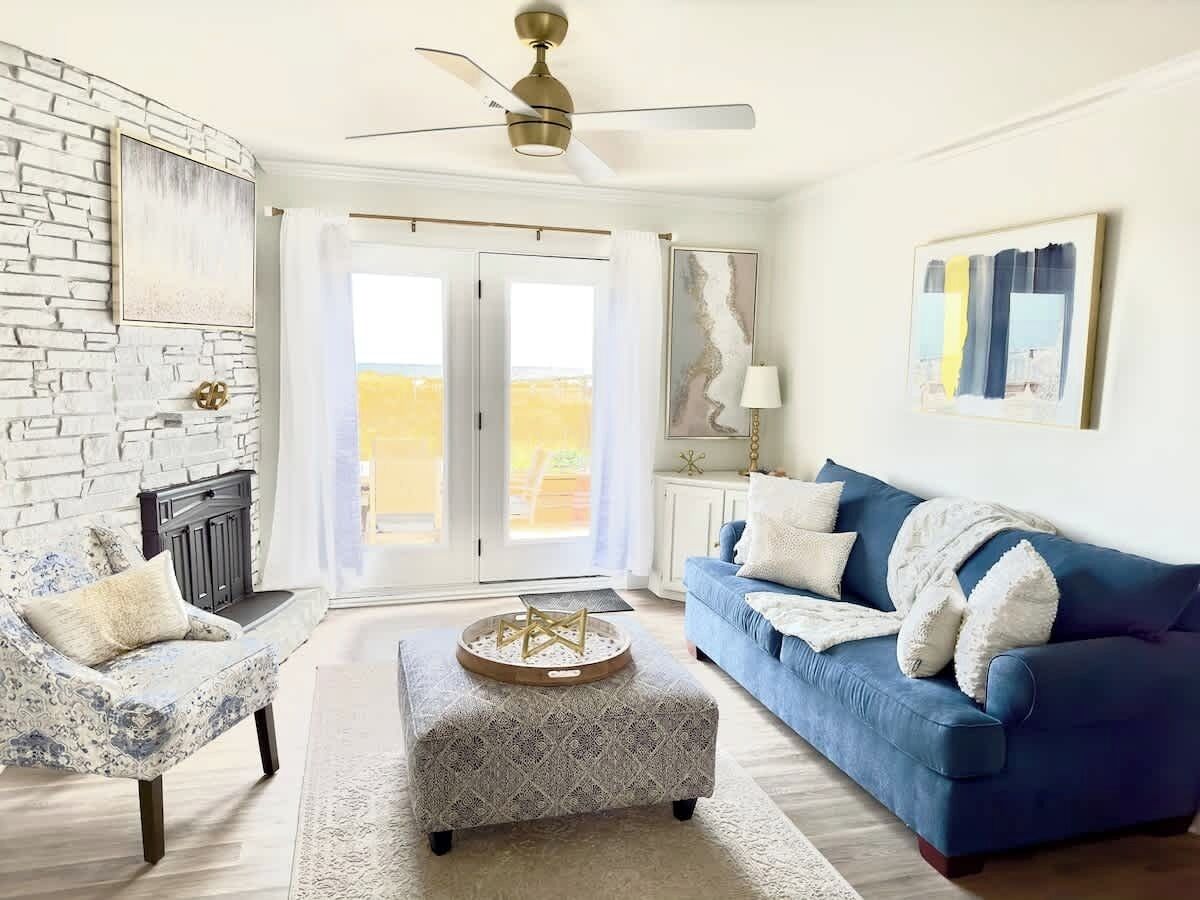 A living room with a blue sofa, patterned armchair, and ottoman, a ceiling fan, and a view through glass doors.