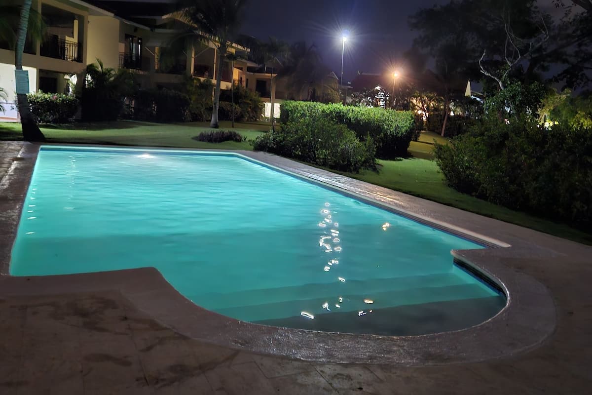 Piscina à noite com vegetação ao redor e um edifício ao fundo.