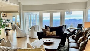 Un vaste salon doté d’une grande fenêtre offrant une vue sur les montagnes, un coin salon confortable avec une table basse, et une cuisine moderne en arrière-plan.