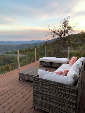 A deck with wicker furniture and cushions, overlooking a mountainous landscape.