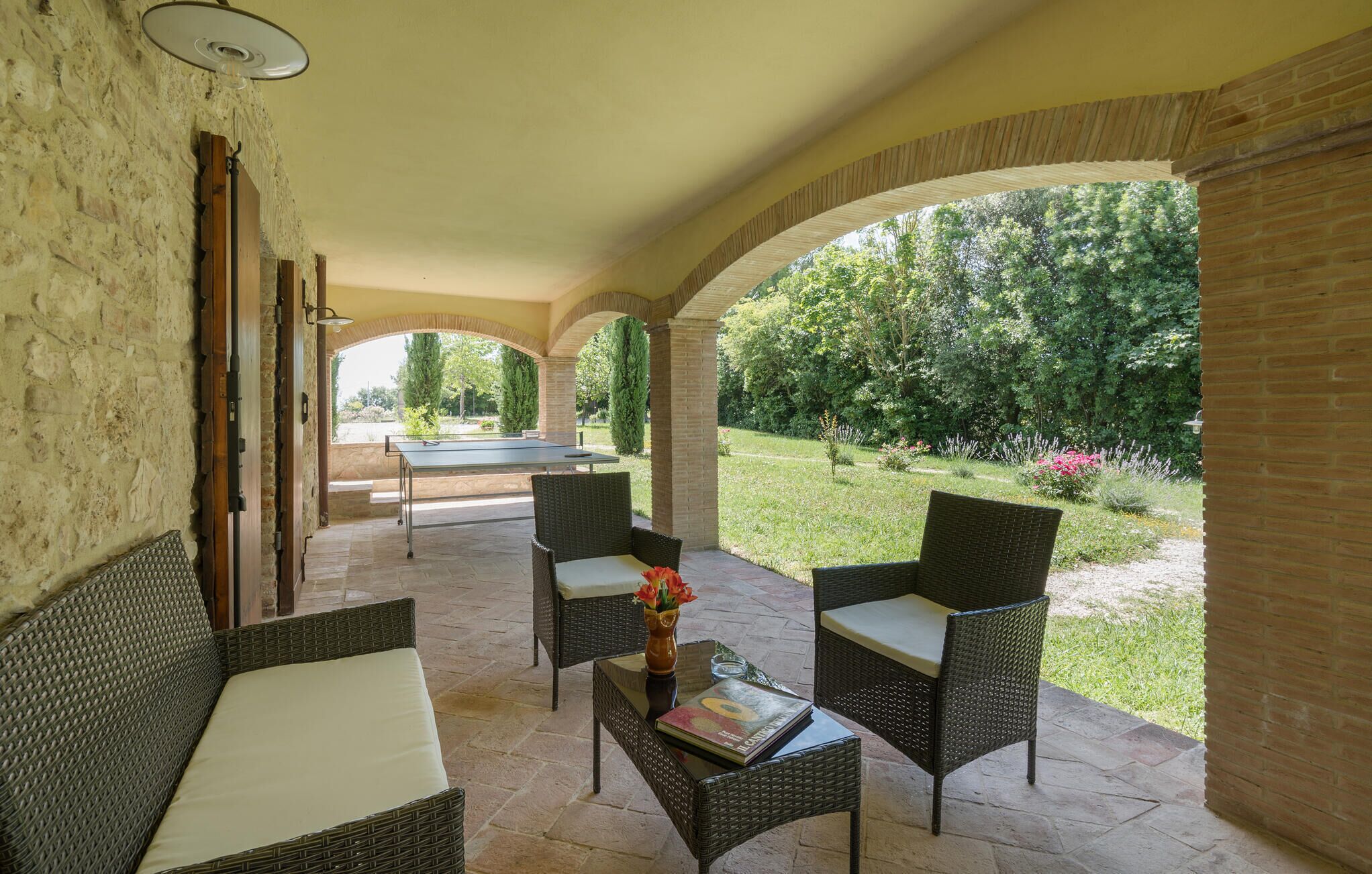 Eine überdachte Terrasse mit Korbmöbeln, einem Tischtennisschläger und Blick auf einen Garten.