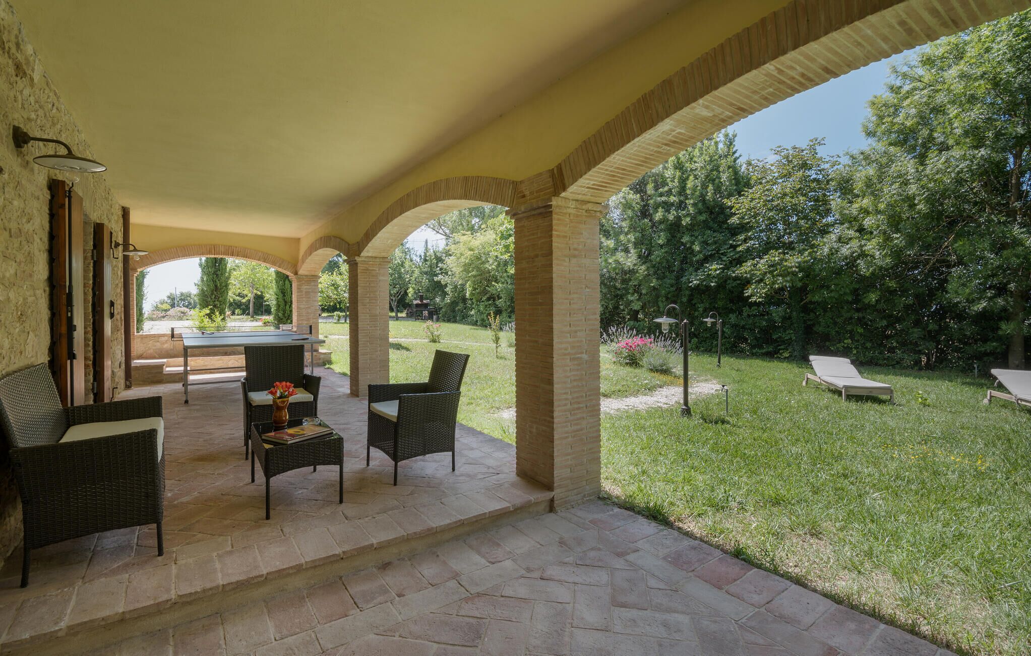 An outdoor patio with a pool table, wicker furniture, and a view of a lush garden.