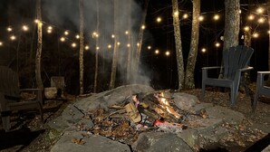 A campfire surrounded by rocks, with two Adirondack chairs and a string of lights in a forest at night.