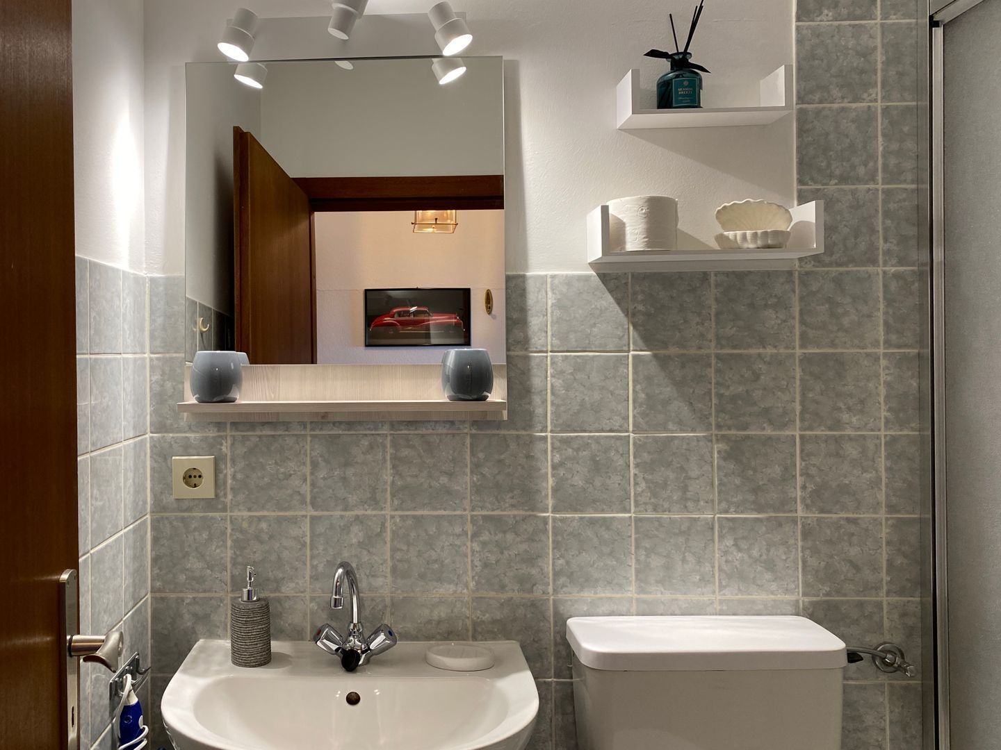 A bathroom with a sink, toilet, mirror, and shelves with toiletries.