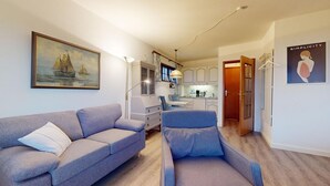 A living room with a sofa, a chair, a painting, a kitchen area, and a door leading to another room.