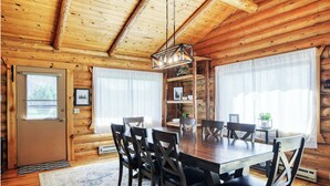 Interior de una cabaña de madera con una mesa de comedor, sillas, una araña y una estantería.