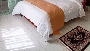 Una cama bien hecha con una colcha blanca y anaranjada, un cabecero rojo y una alfombra estampada sobre un suelo de baldosas.