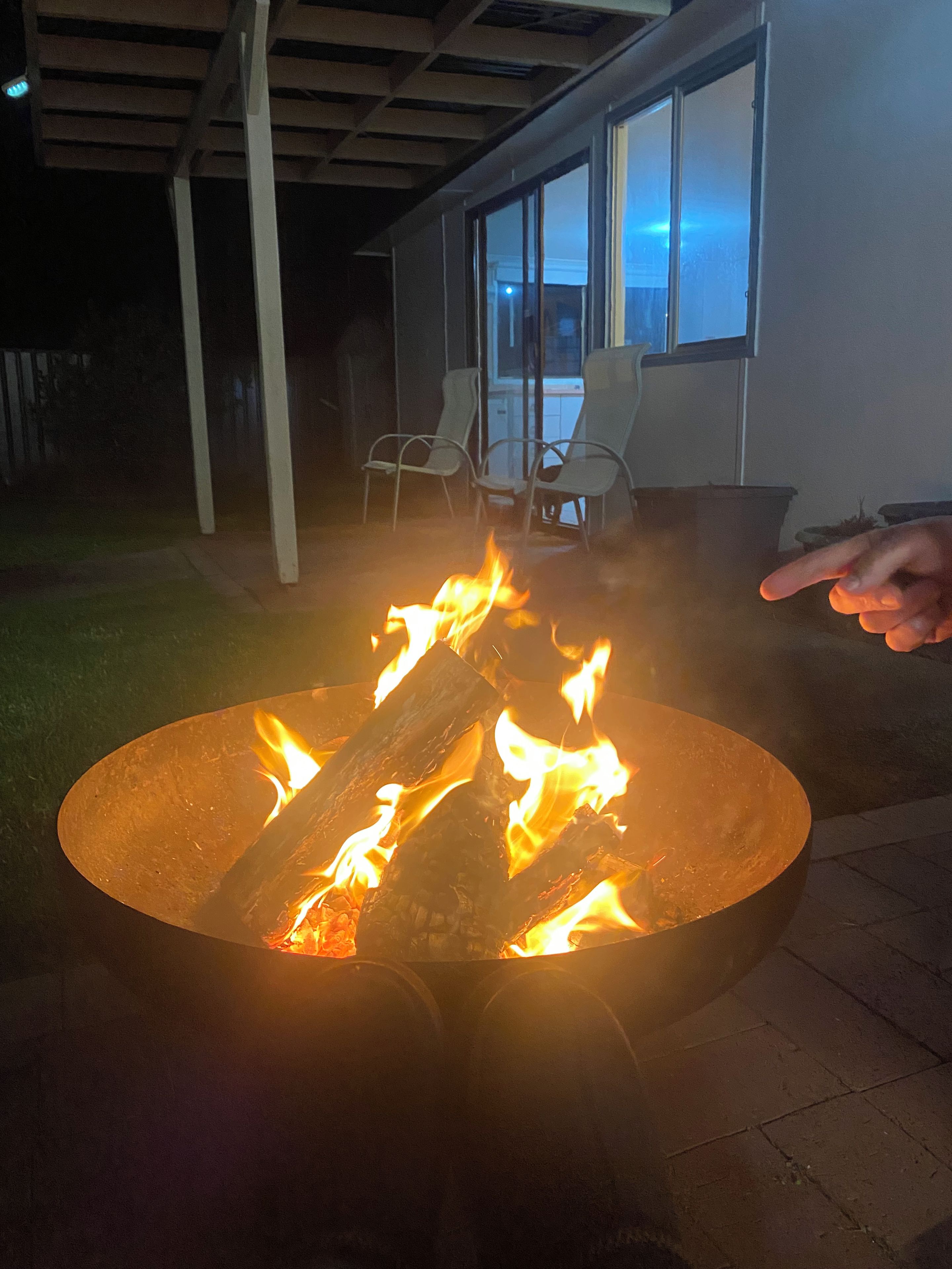 A fire pit with logs burning, surrounded by outdoor furniture and a house in the background.