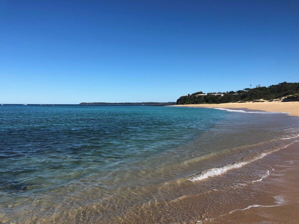 A beach with clear water, sandy shore, and a hill with buildings.