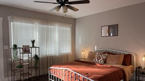 A bedroom with a bed, a ceiling fan, a window with sheer curtains, a bedside table with a lamp, and a wall-mounted picture.