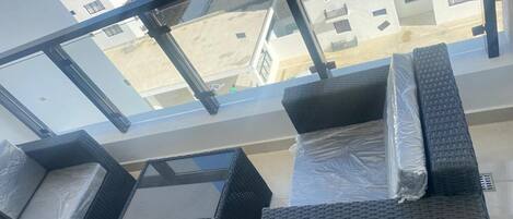 Un balcon doté de meubles en osier, d’une rambarde en verre et offrant une vue sur les bâtiments en arrière-plan.