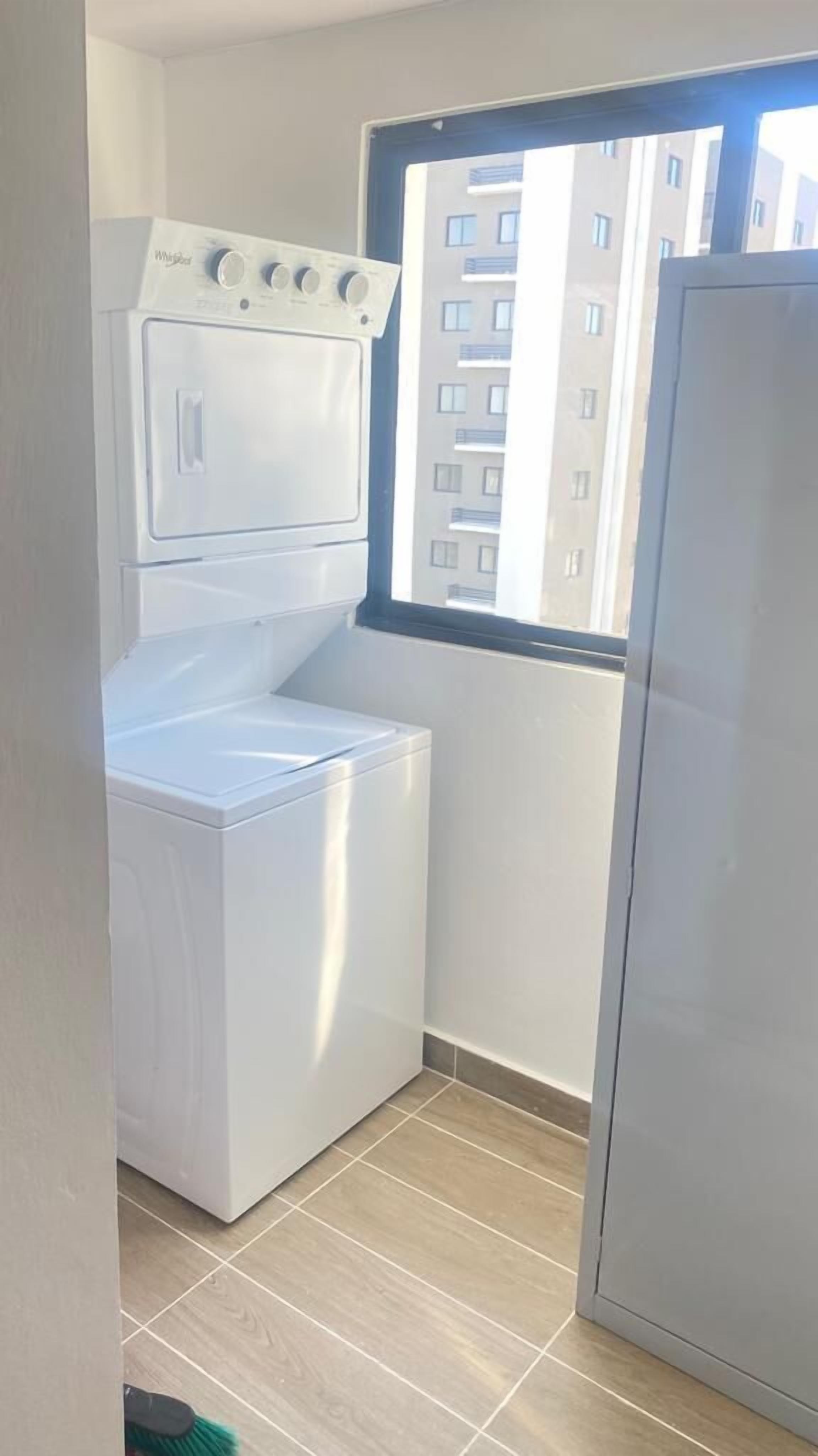 Een wasserette met een wasmachine en droger, een raam en uitzicht op een gebouw.