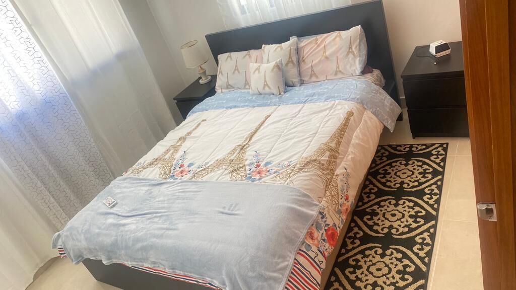 A neatly made bed with a floral and Eiffel Tower themed duvet, a bedside table with a lamp, and a patterned rug on the floor.
