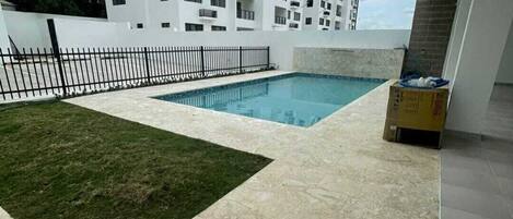 A rooftop pool area with a tiled floor, a grassy patch, and a white building in the background.
