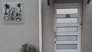Une porte blanche portant le numéro 23B, une enseigne indiquant « Palm Cottage », et un bac à fleurs avec des plantes placé à côté de la porte.