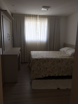 Quarto com cama, mesa de cabeceira, guarda-roupa e janela com cortinas.