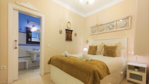 A bedroom with a bed, bedside table, and a nightstand. There is a bathroom visible through an open door.