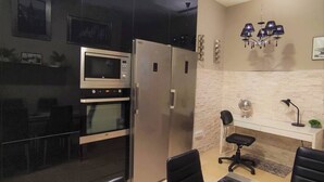 A modern kitchen with a built-in oven, microwave, and stainless steel refrigerator. There is a dining area with a table and chairs, and a desk with a chair and a lamp.