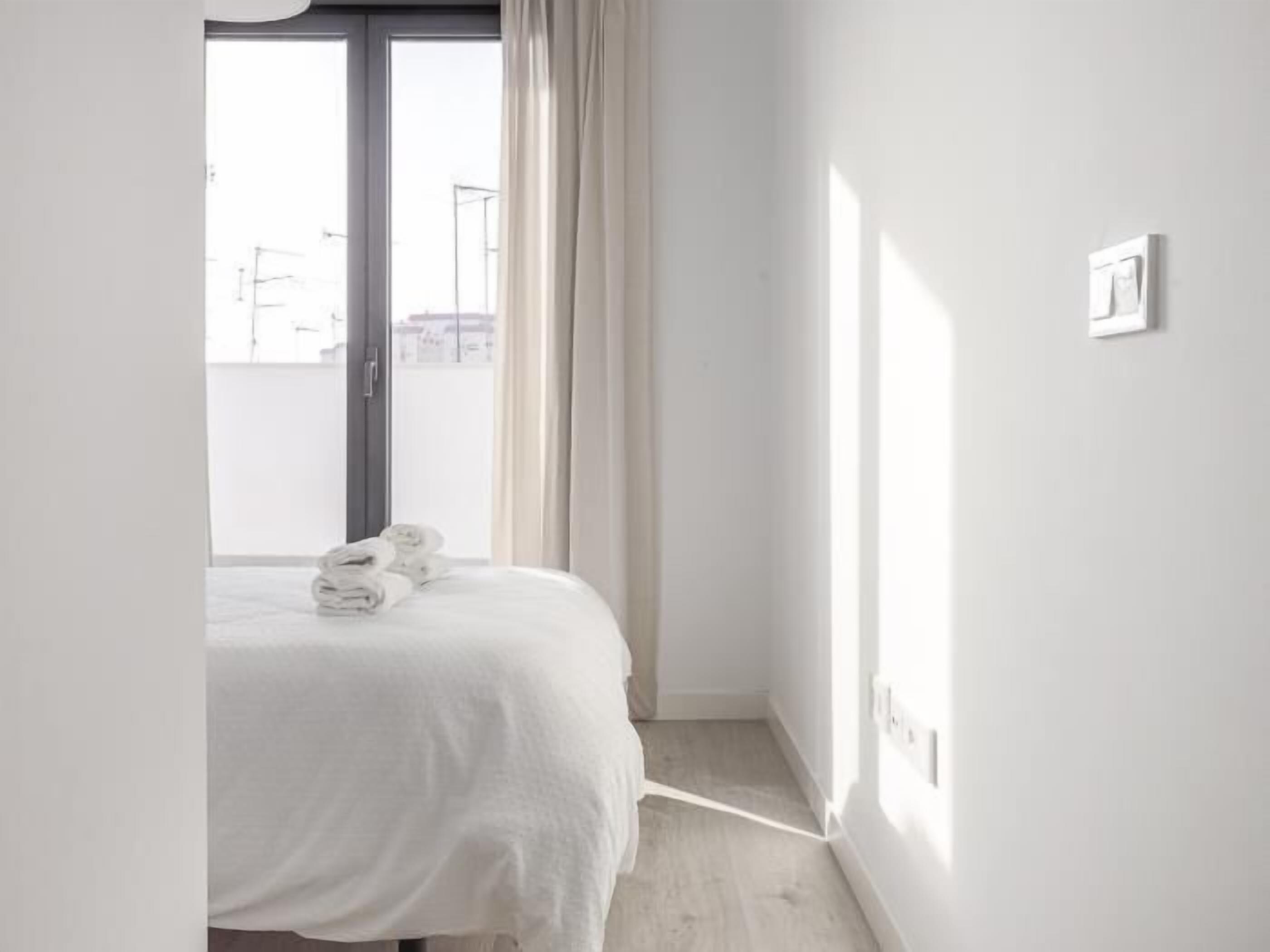 A minimalist hotel room with a bed, a window with curtains, and a view of the outside.
